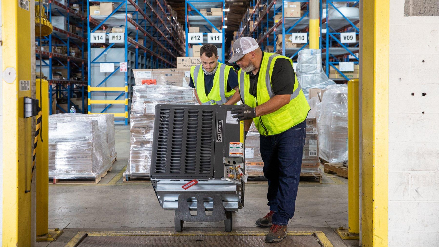 Two Ferguson associates wearing reflective vests push an HVAC condenser on a dolly at a warehouse location.