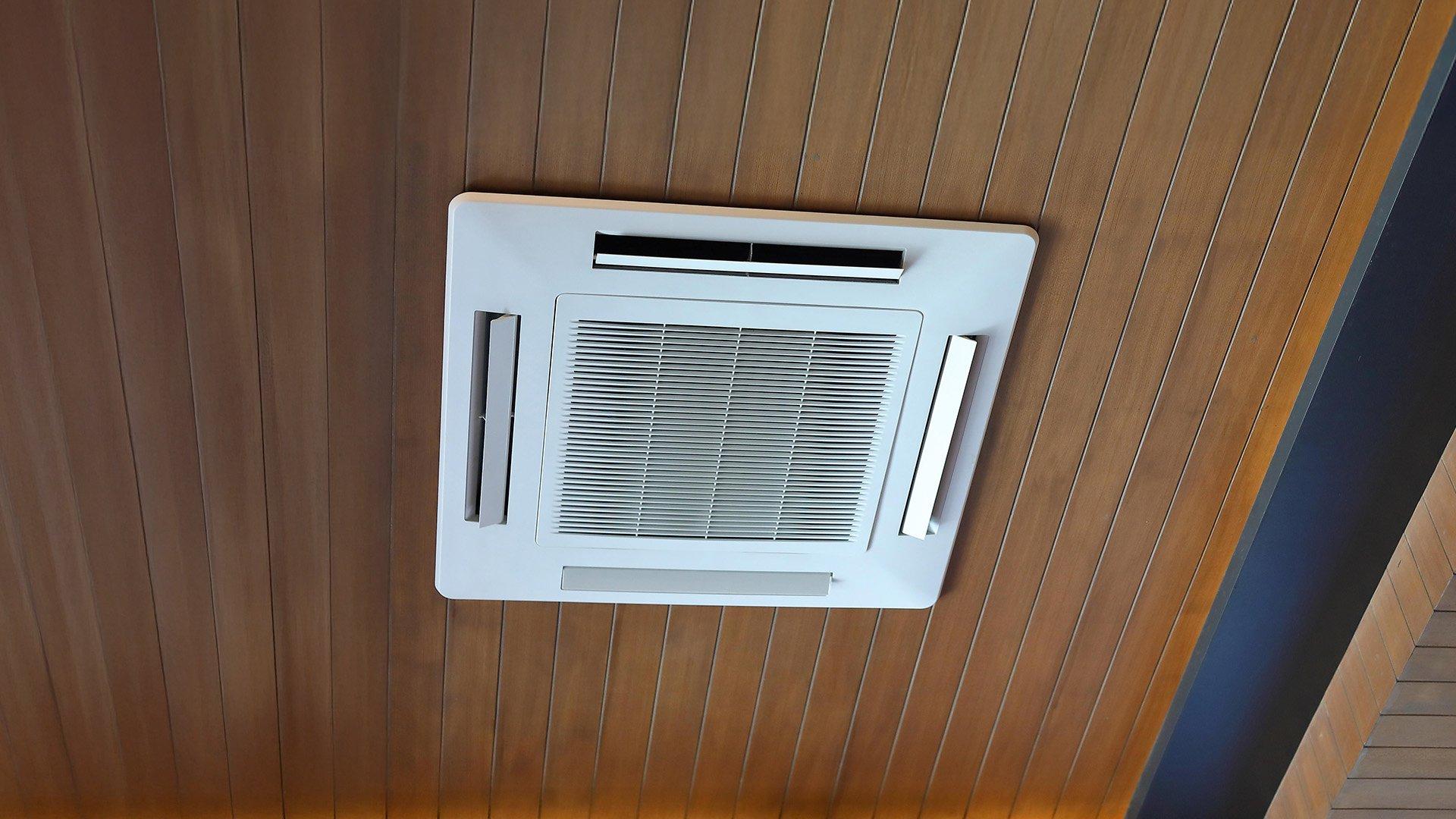 A mini-split air conditioner cooling cassette is installed on a wooden ceiling.