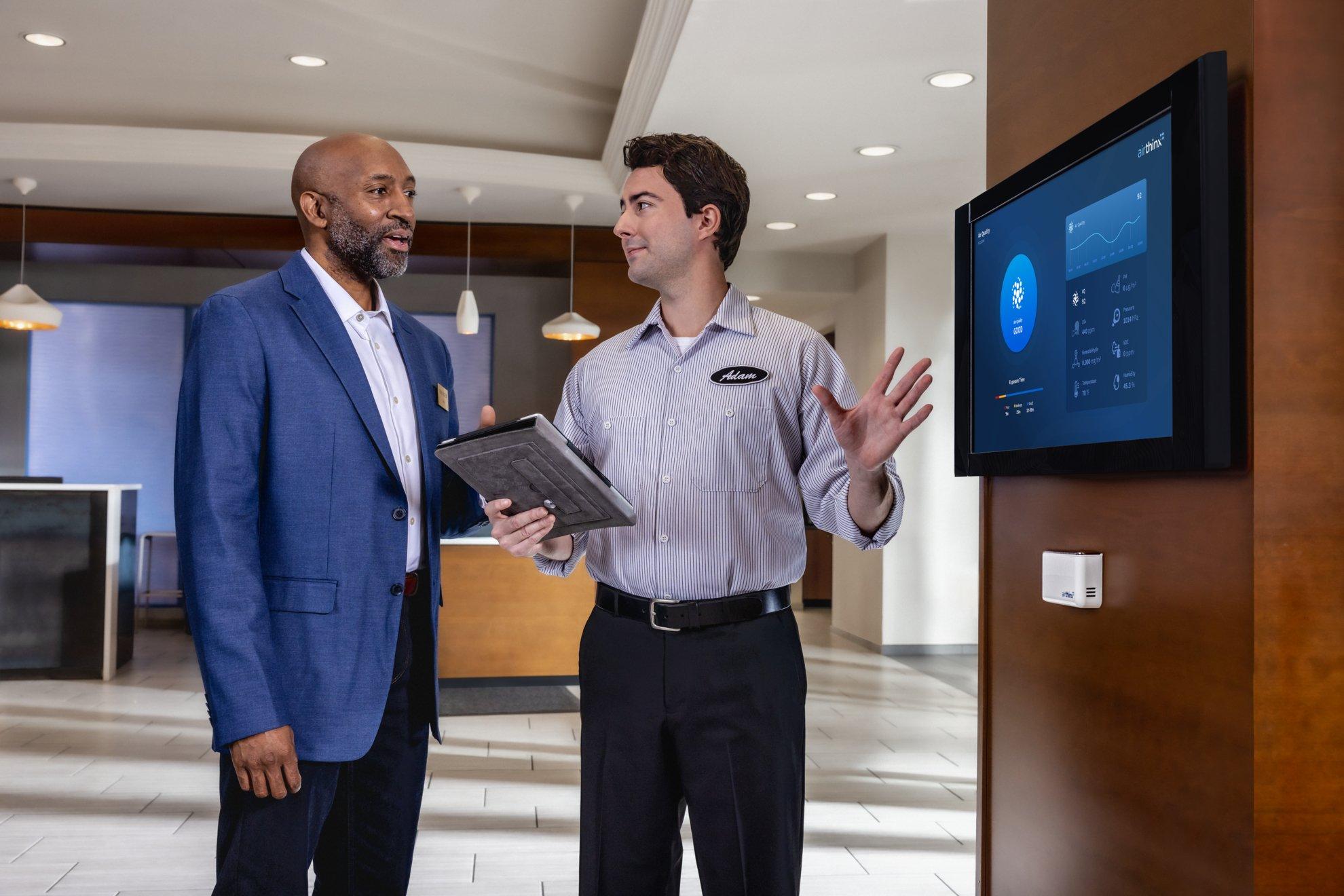 In a hotel lobby, an HVAC technician speaks with a hotel executive in front of an IAQ monitoring screen.