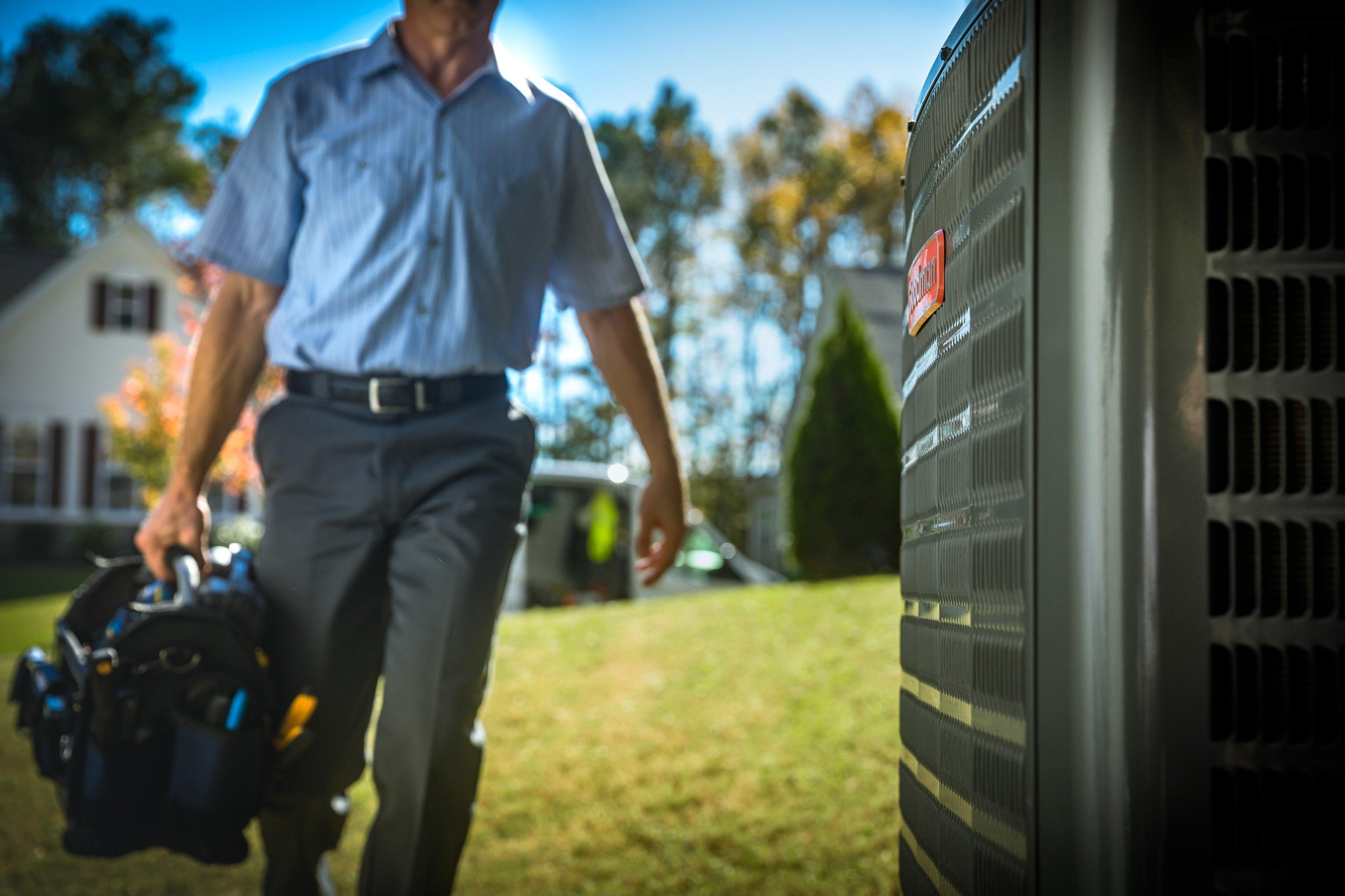 An HVAC technician carrying his toolkit walks across a residential lawn toward an air conditioner condenser.