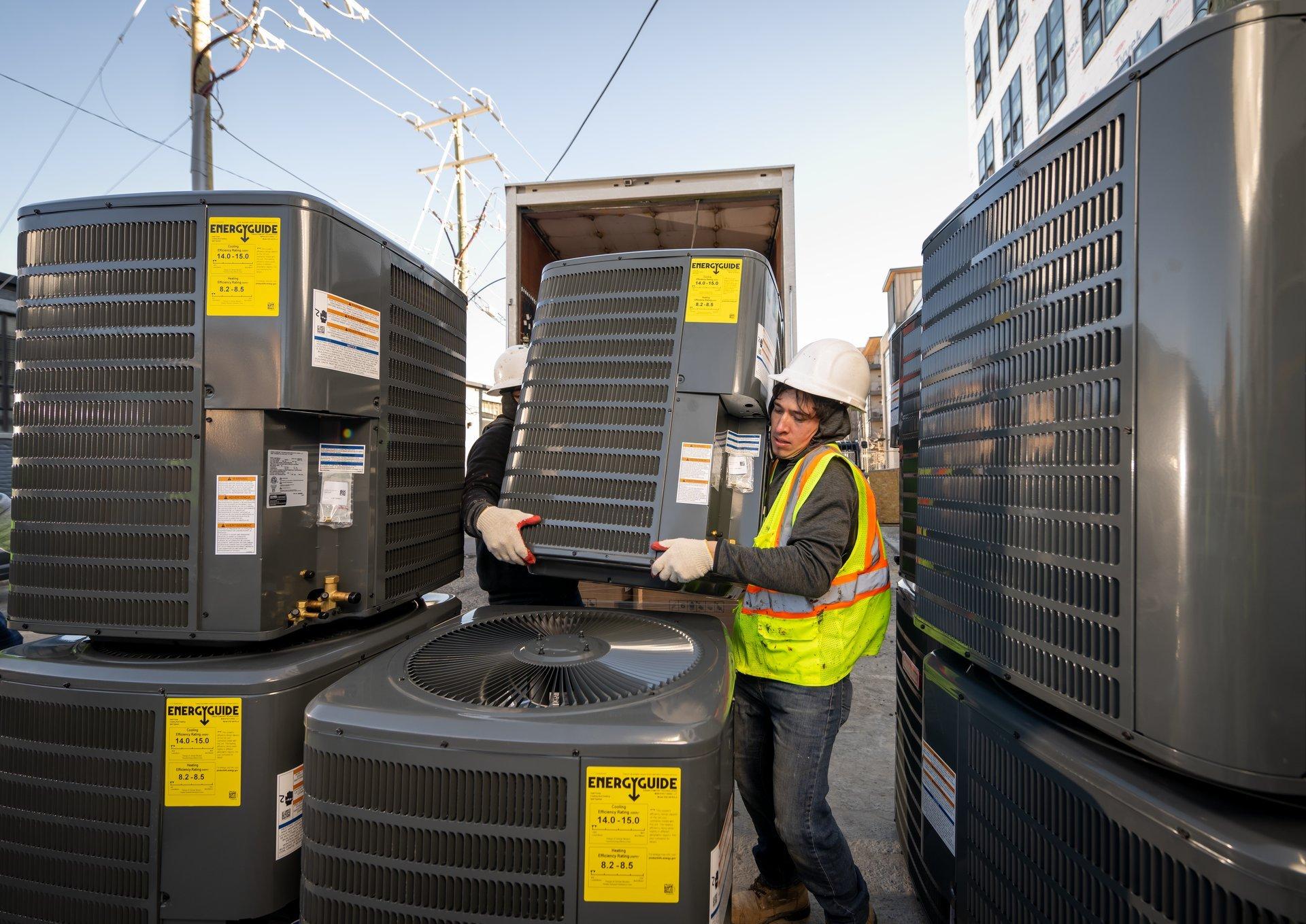 Two workers wearing PPE carry an air conditioner condenser.