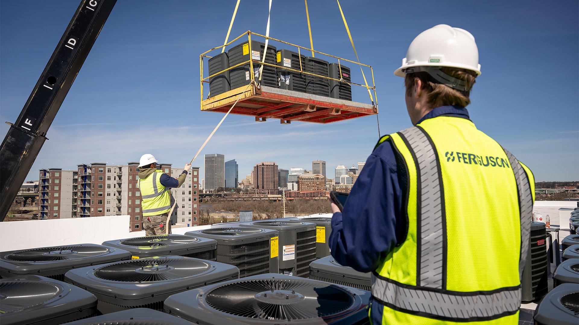 A crane lowers an HVAC condenser onto the roof of a building, while two Ferguson associates guide it into place among two rows of condensers.