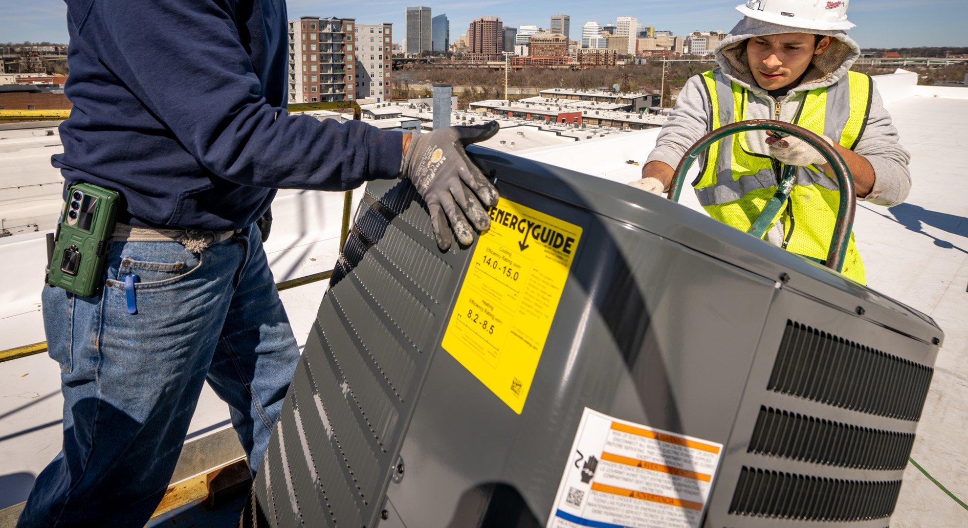 Two HVAC technicians use a dolly to haul a condenser on a rooftop.