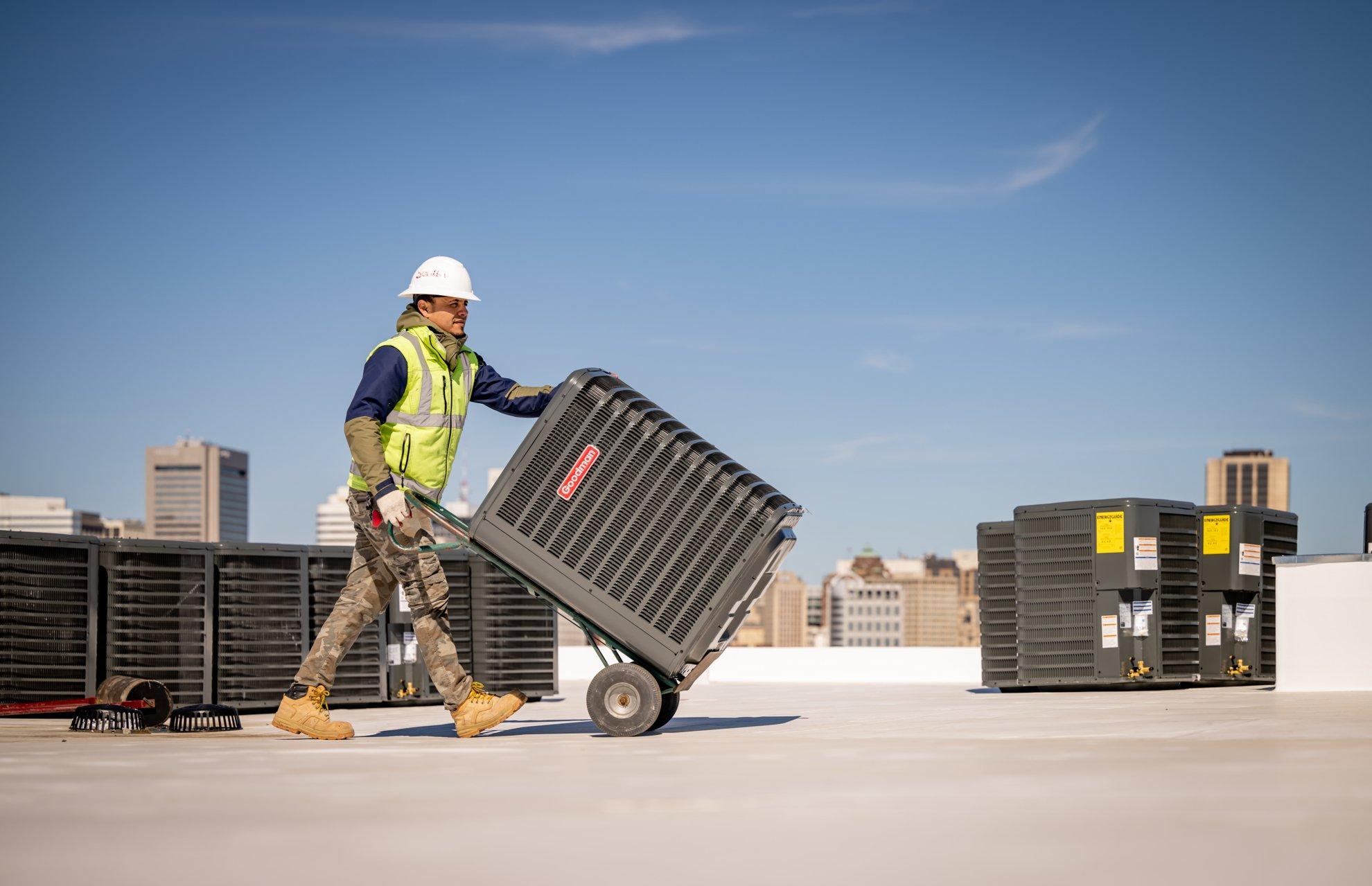 An HVAC technician uses a dolly to push an air conditioner condenser across the roof of a commercial building.