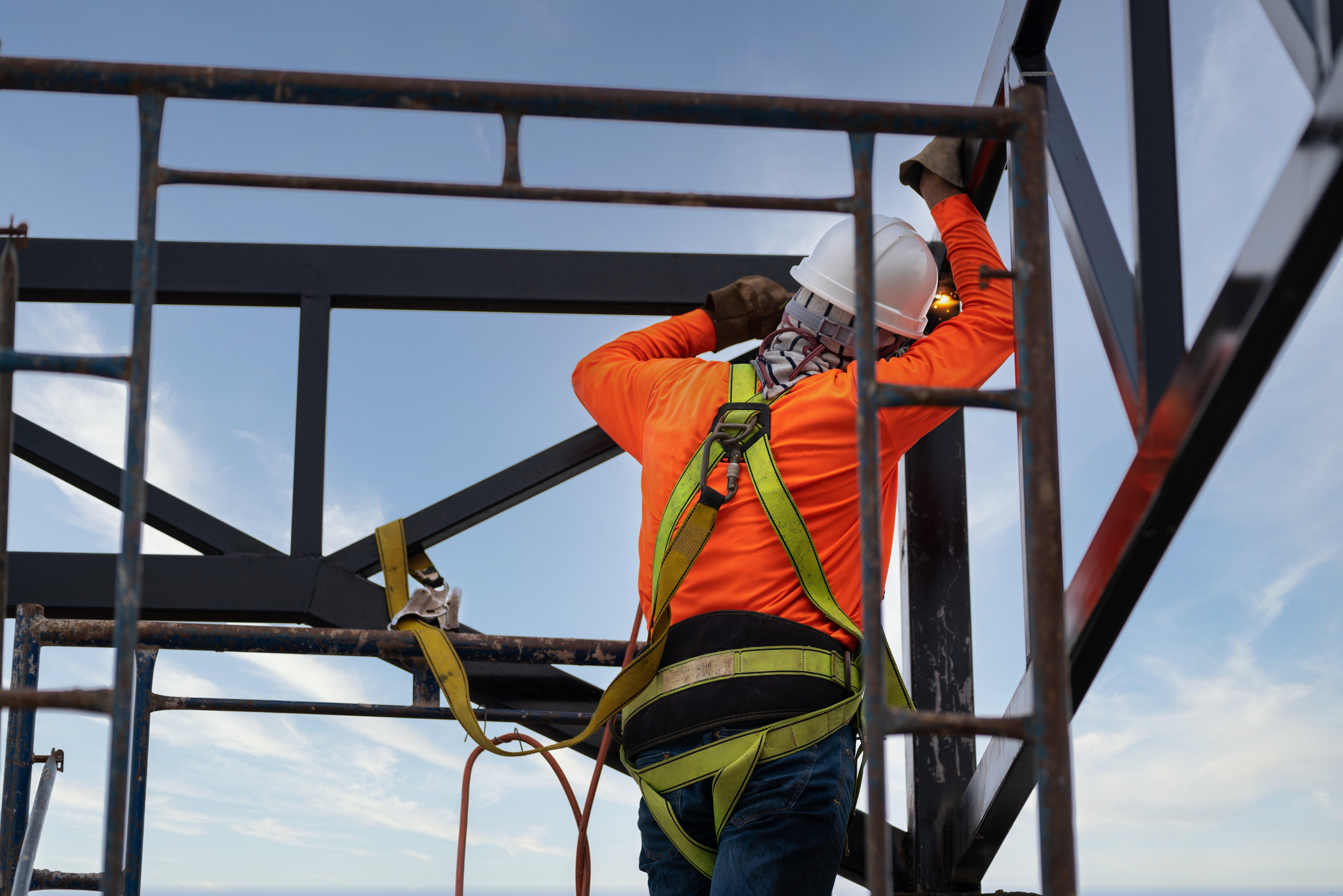 A worker wearing reflective clothing uses a fall arrestor device and safety body harness while welding steel on a construction site.