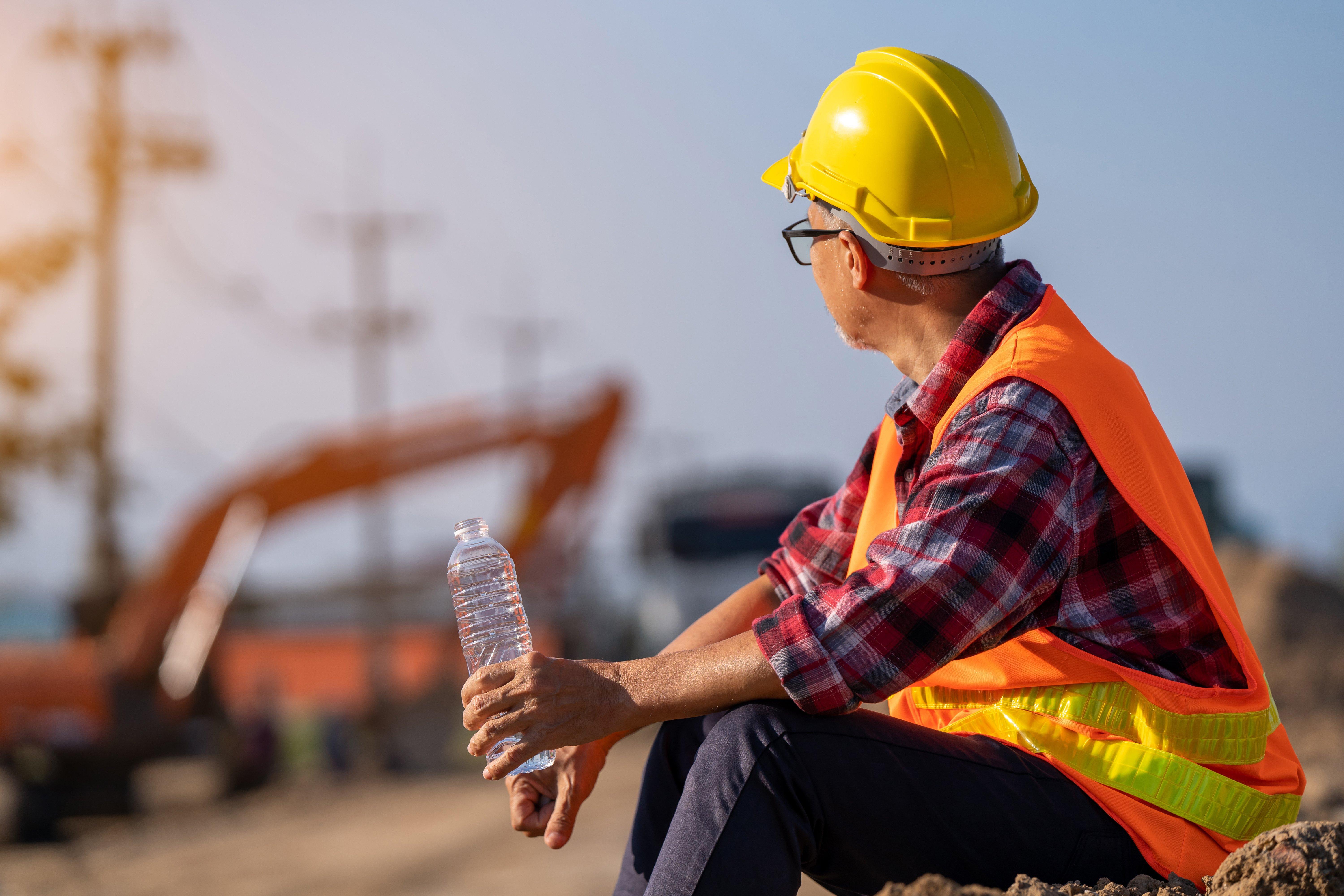 A construction worker wearing a hard hat and reflective vest holds an open water bottle while sitting on a construction jobsite.