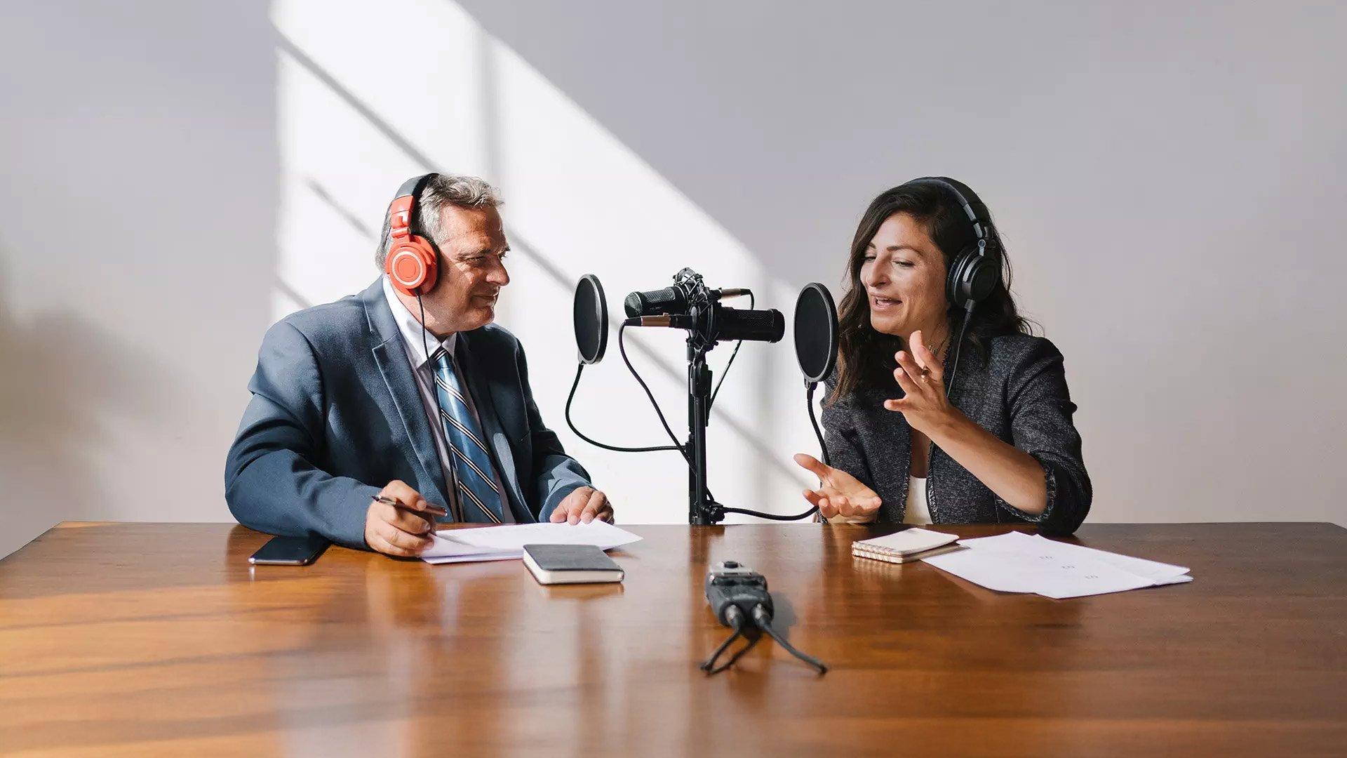 Male and female podcast hosts sit at table with headphones and microphone.