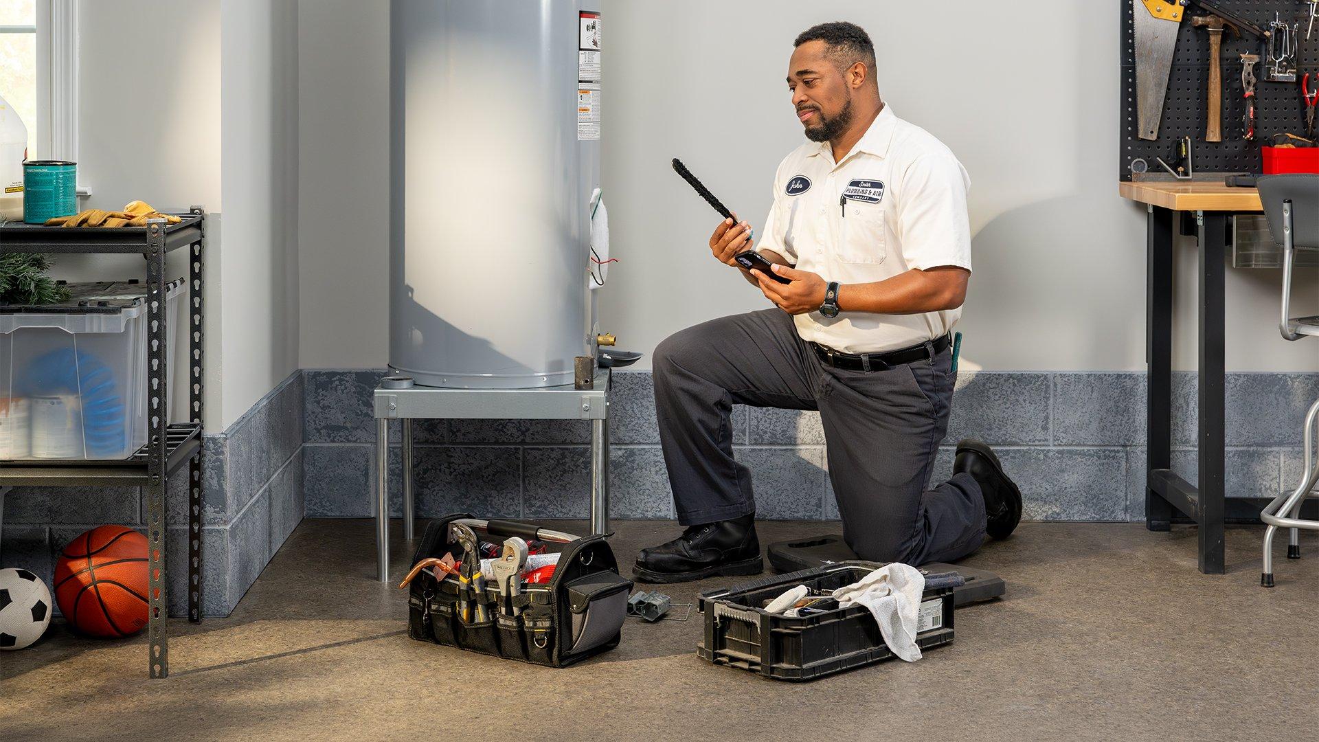 Plumbing contractor kneels in front of installed water heater in residential garage with toolbox in front of him.