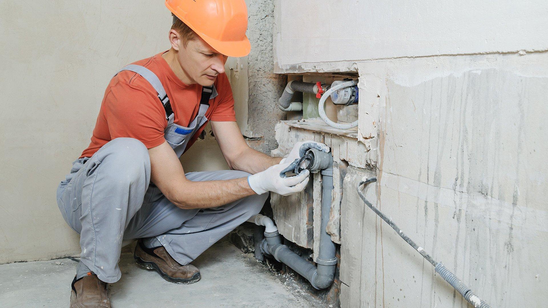 A worker in a hardhat repairs pipe in the wall.