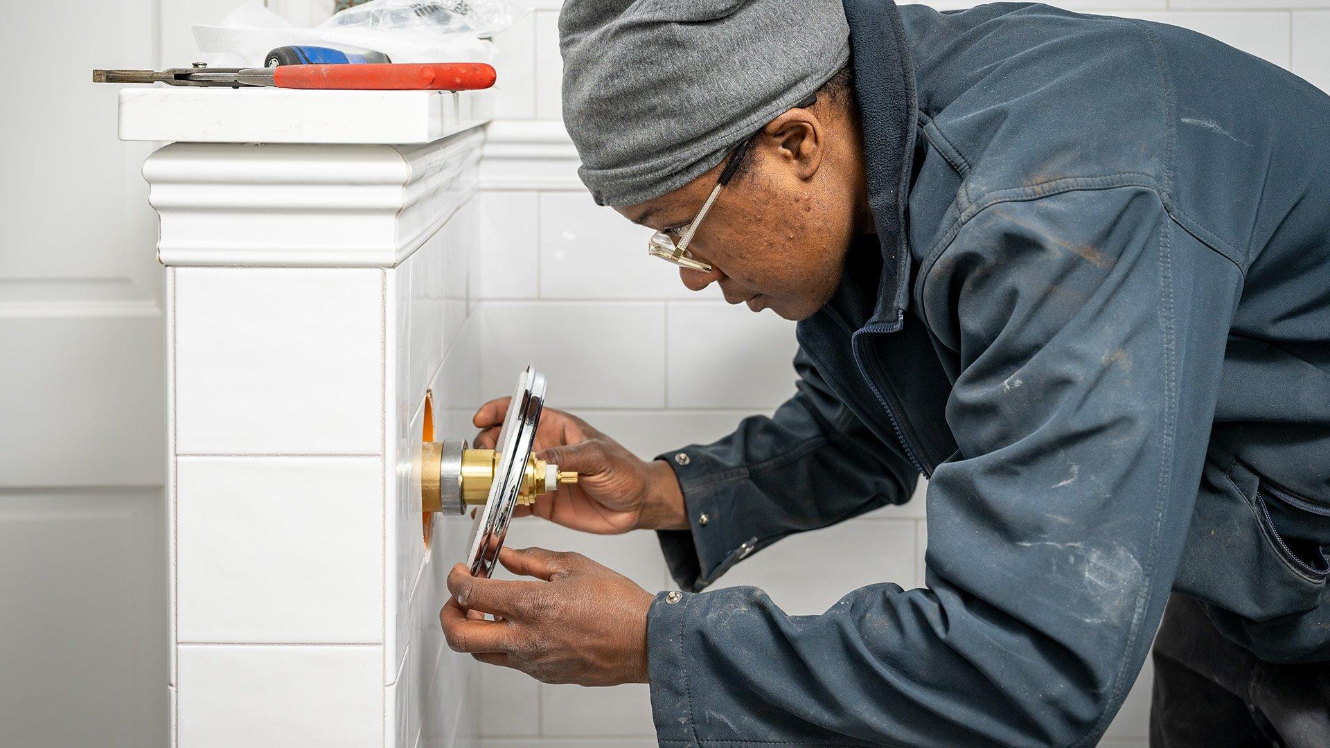 Worker stands in unfinished bathroom and installs white tile in shower.