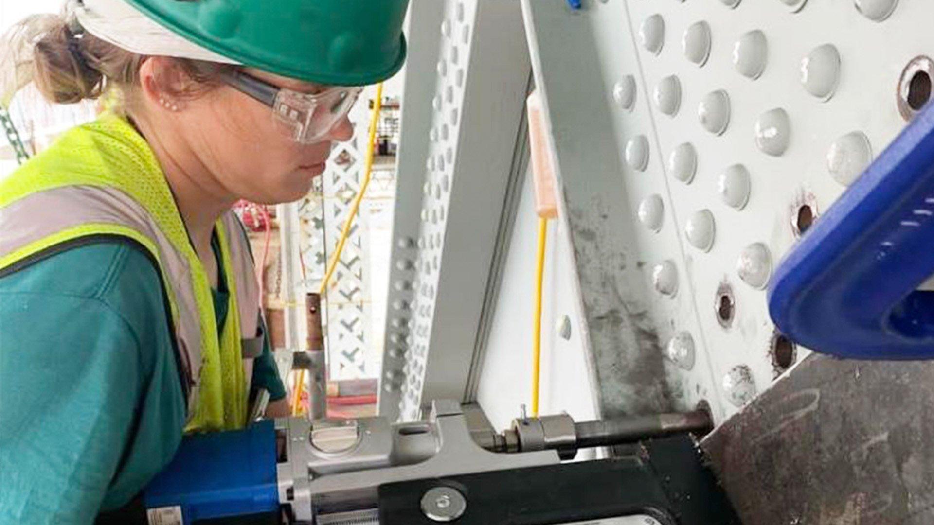 Ali G wears a green hard hat and safety glasses while using a large portable magnetic drill on a bridge.