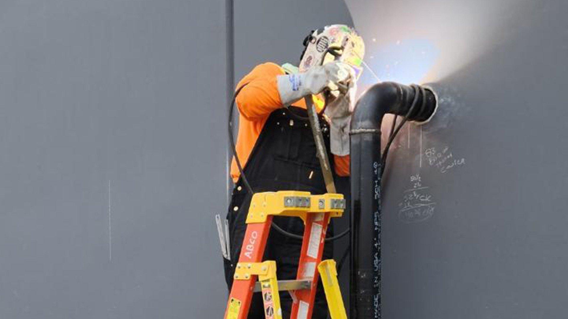 Emily S, wearing a welding helmet and protective gloves, stands on a ladder and welds a black pipe next to a gray wall.