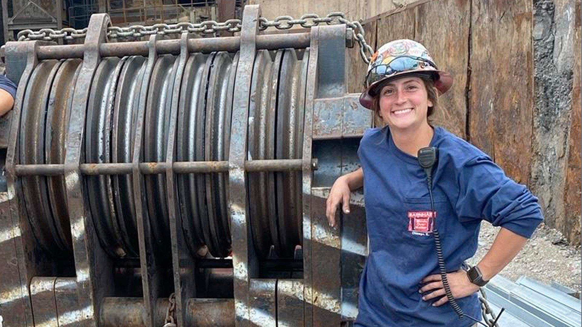 Erin M wears a hard hat and smiles next to an industrial winch with chain on a jobsite.