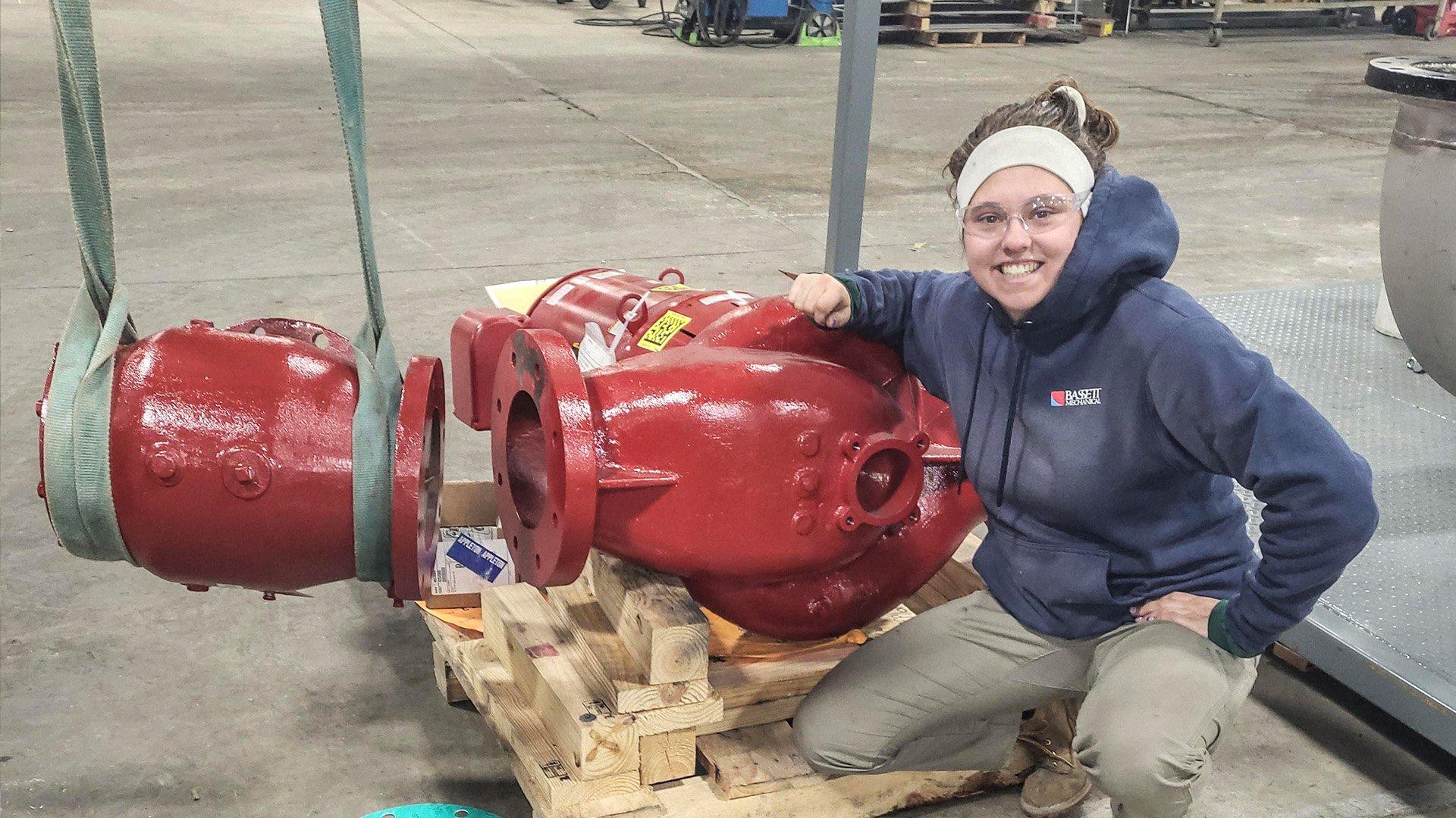 Grace T smiles next to red underground pipe connectors in a warehouse facility.