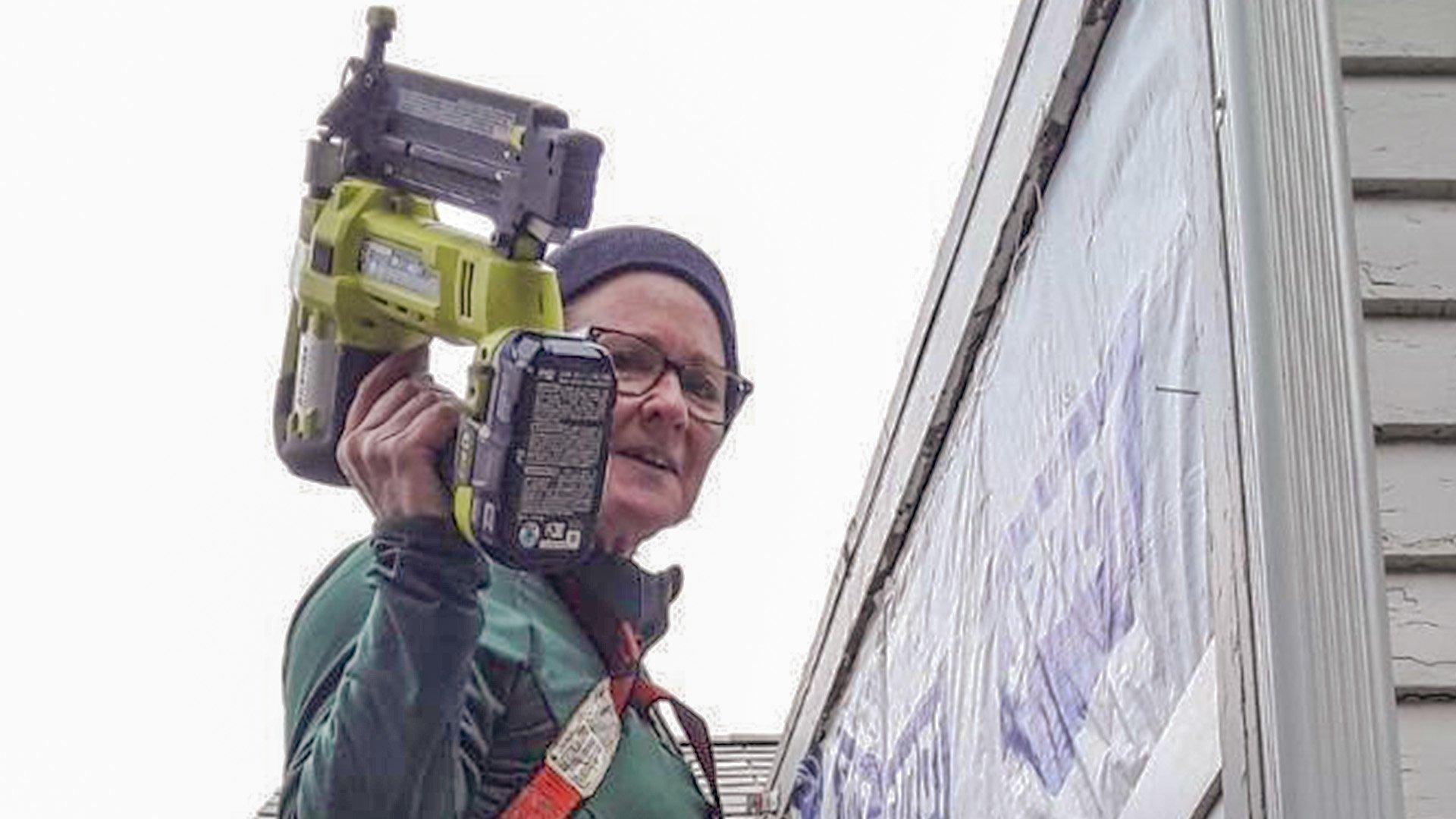 Karen L wears fall protection gear while standing on a residential roof. She holds a level and a nail gun to work on siding.