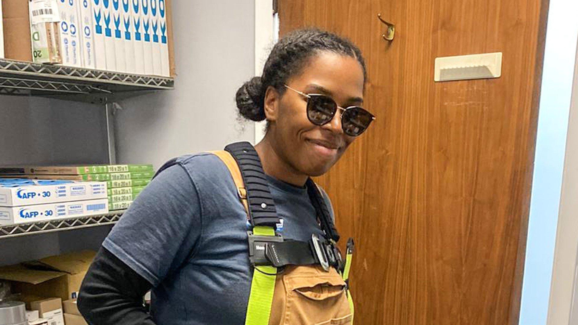 Shaquia J wears sunglasses, yellow fall protection gear and a smile at work, with HVAC supplies on shelves in the background.