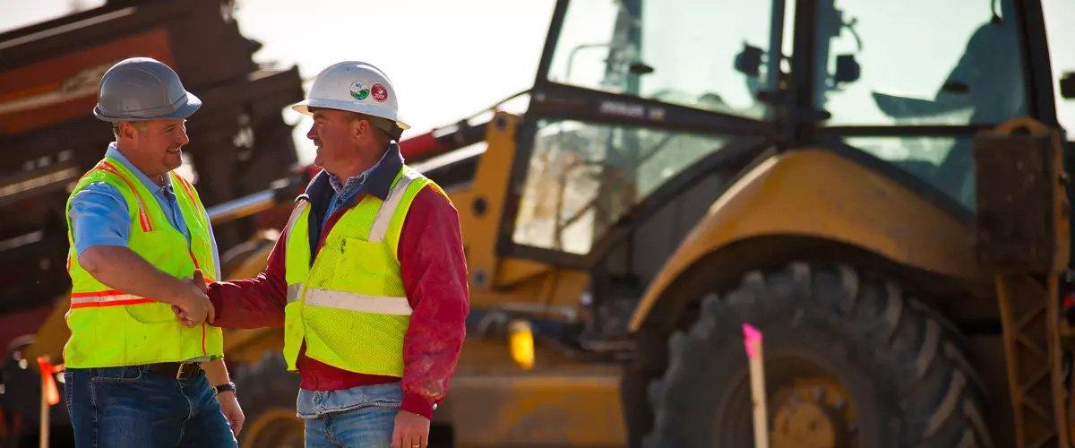 Two contractors wearing hard hats and reflective vests shake hands on a jobsite, with an excavator behind them.