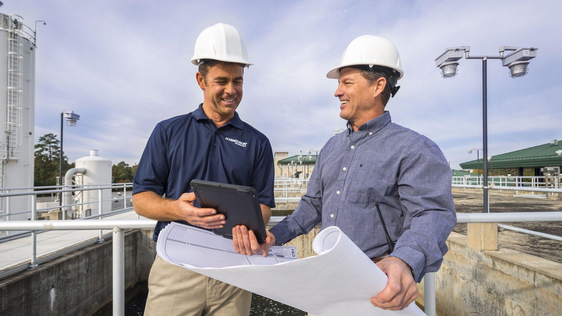 Ferguson associate looks over blueprints with contractor while holding tablet and wearing white hardhats. 