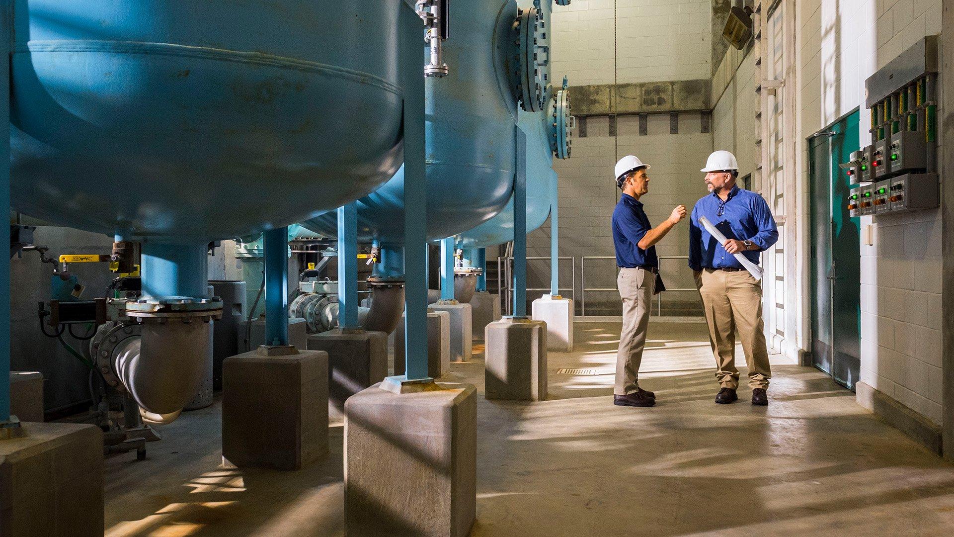Two associates with hardhats stand inside water treatment facility and carry on a conversation. 