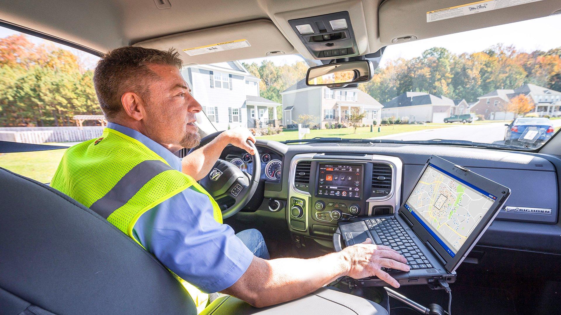 A man wearing a reflective vest looks at a map on an open laptop while he is parked in his work truck in a residential neighborhood.