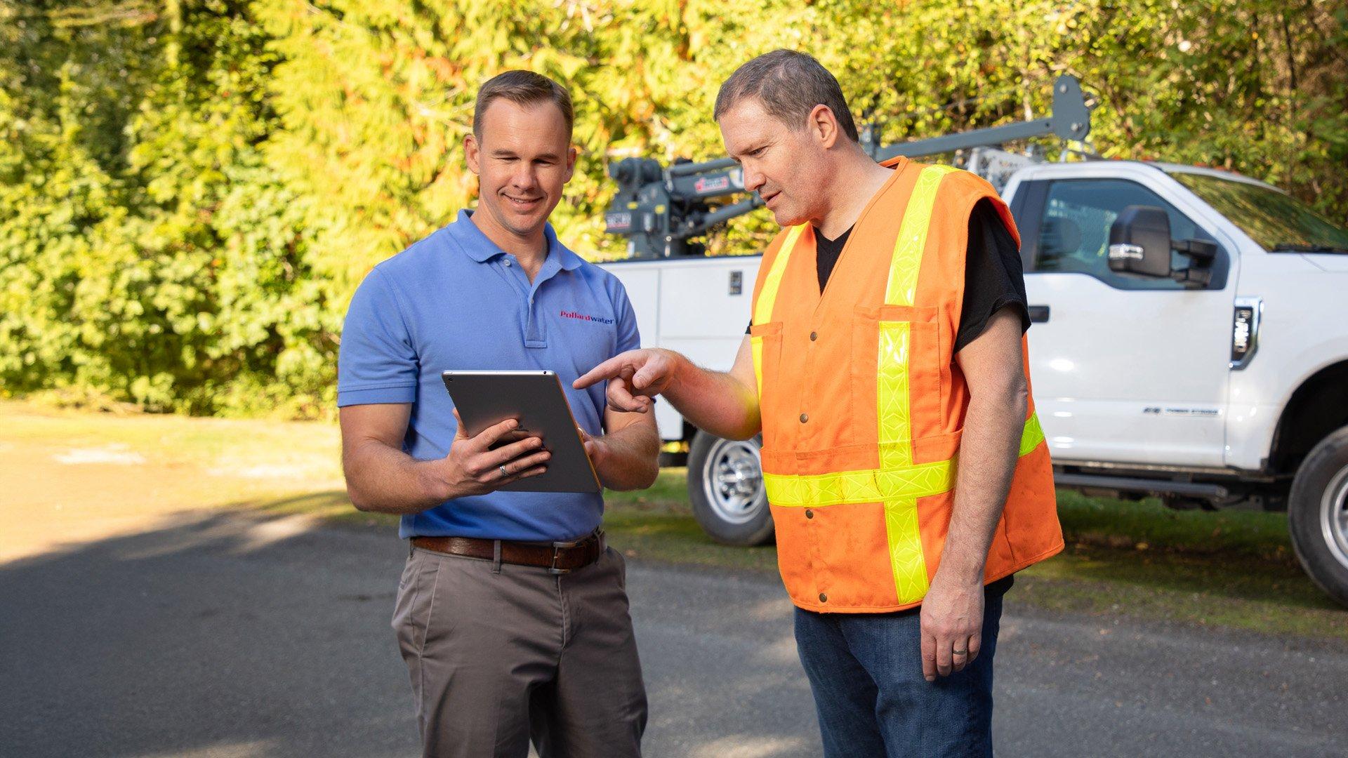 Two men stand outside in front of a white truck and look at a tablet.