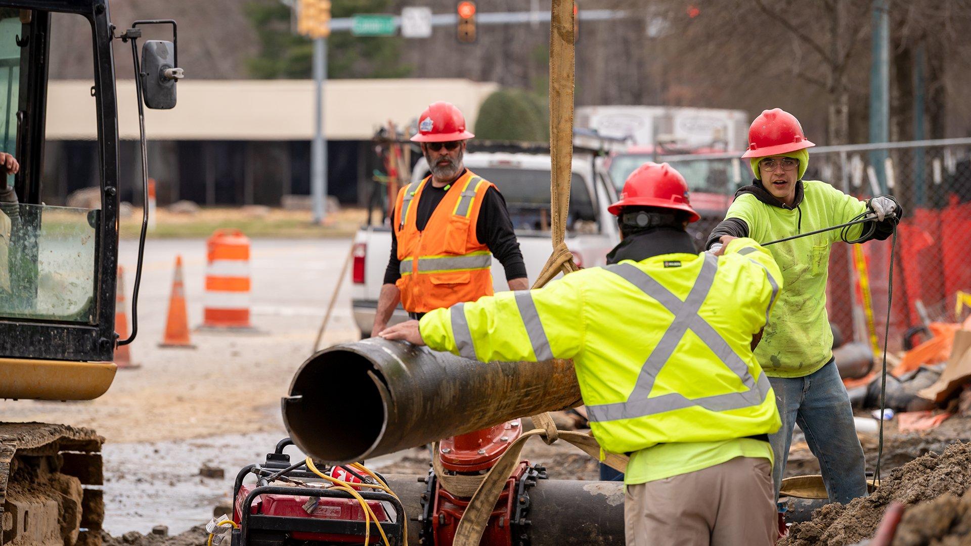 Two workers wearing hard hats and reflective gear maneuver an underground pipe on a jobsite by a public road while a supervisor looks on in the background.