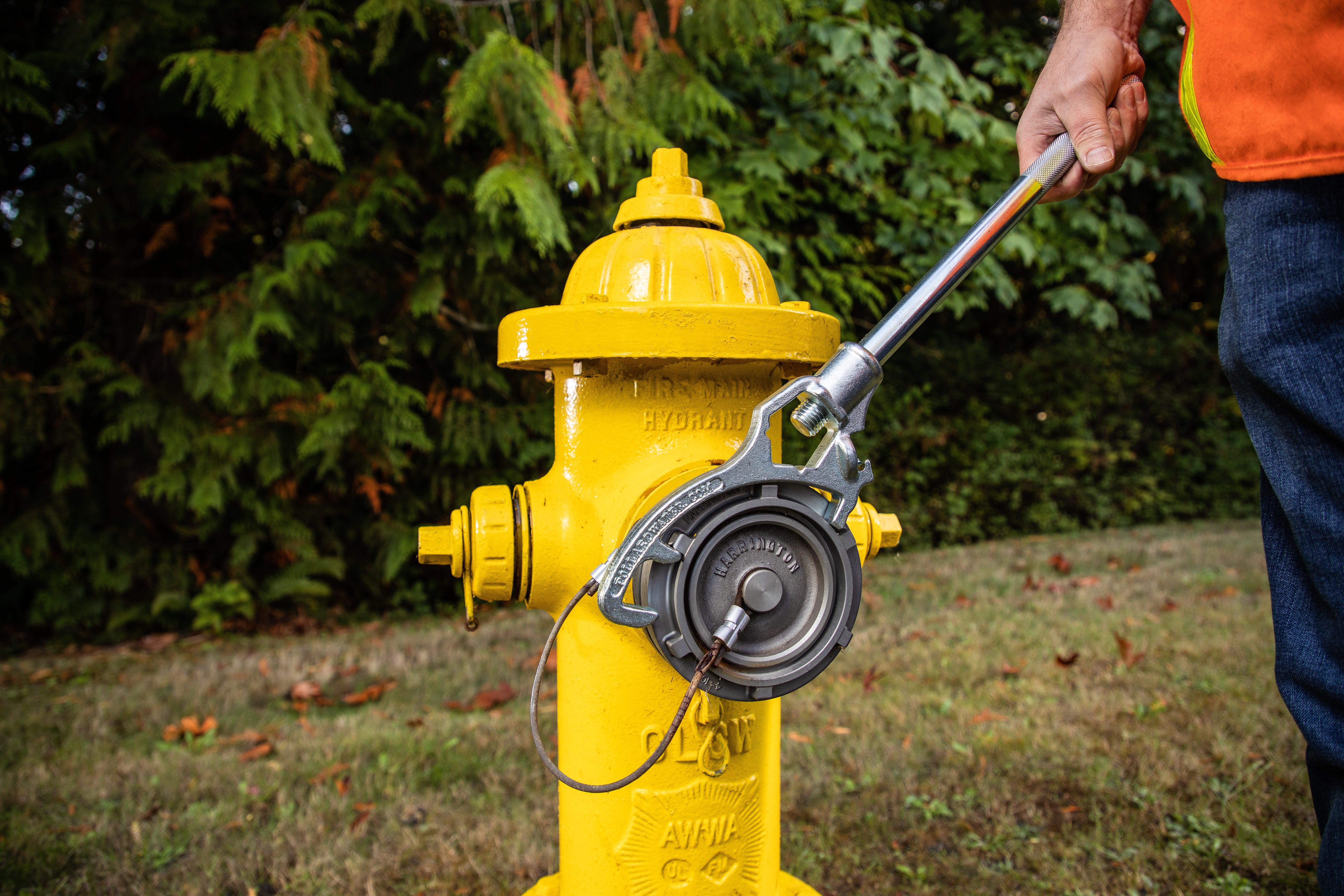 A worker wearing an orange reflective vest uses a wrench to tighten a yellow fire hydrant.