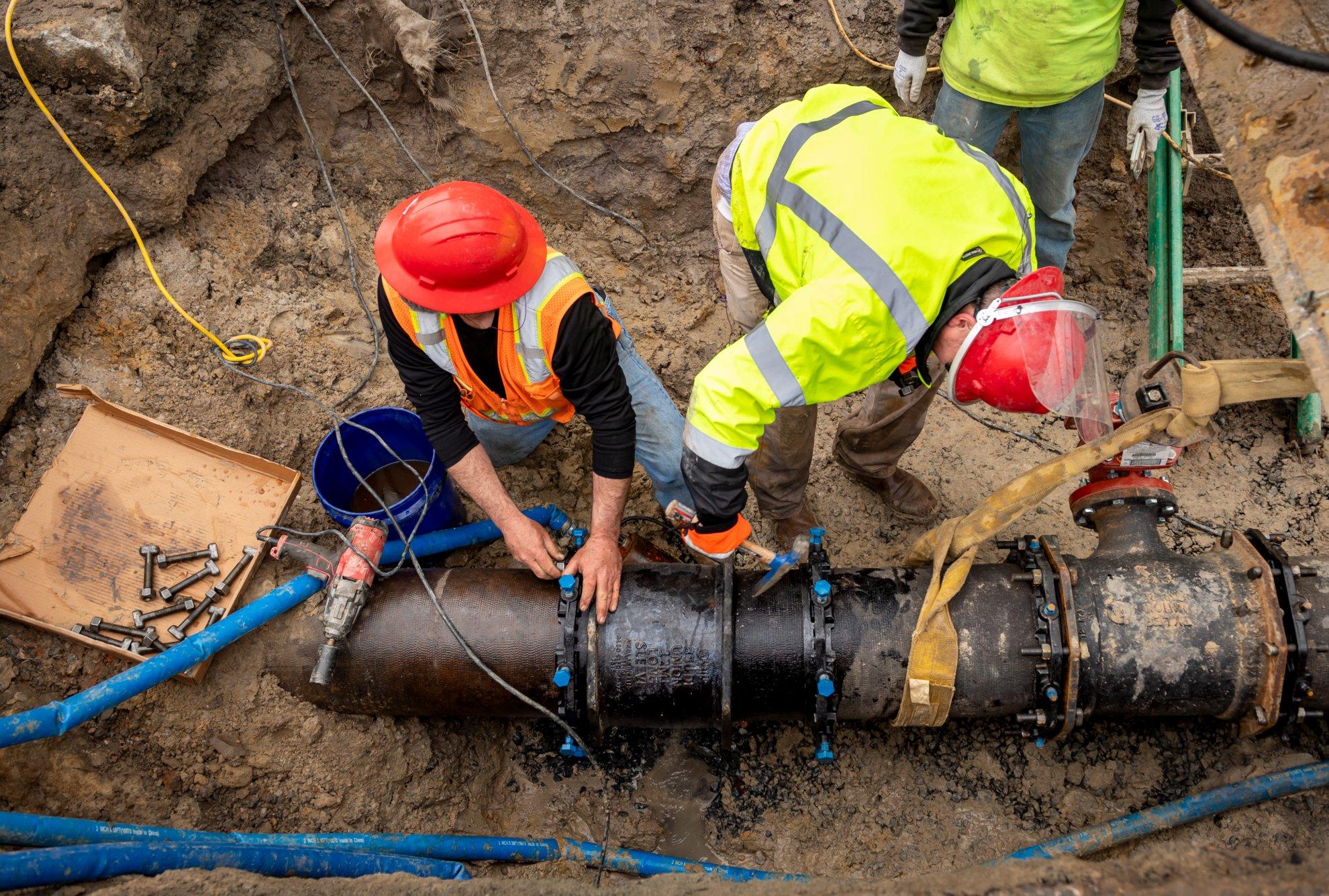 Three workers in a trench work on an underground utility pipe.