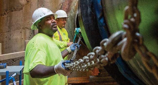 Two workers in hard hats and reflective gear maneuver an industrial pipe into place using chains.