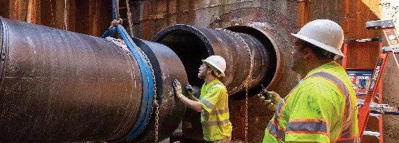 Two workers wearing white hard hats and yellow reflective vests guide the connection of two large underground utility pipes.