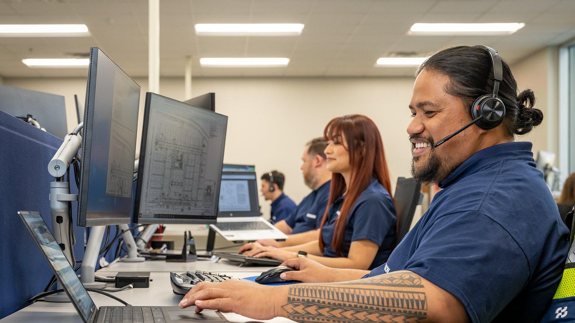 In a call center, a smiling Ferguson associate speaks into a headset while reviewing blueprints on his desk monitor.