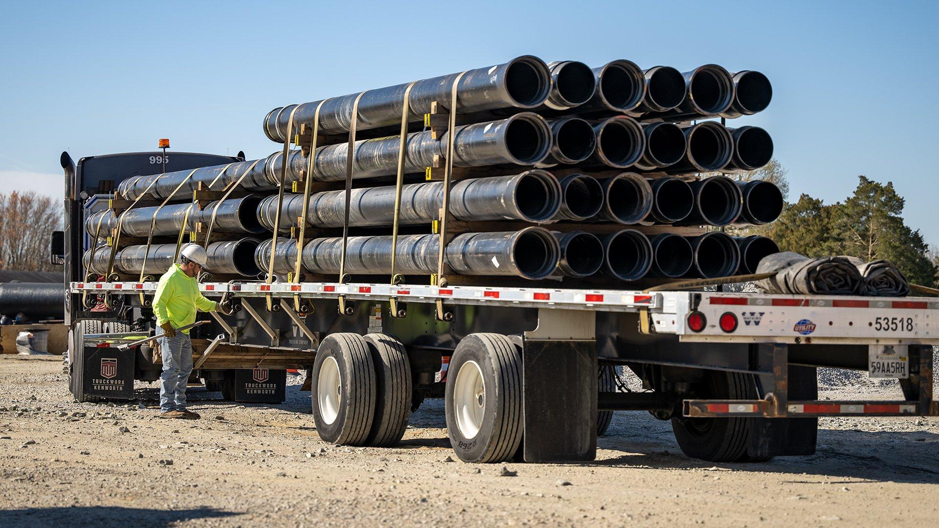 A man in PPE straps down stacks of pipe on an eighteen wheeler.