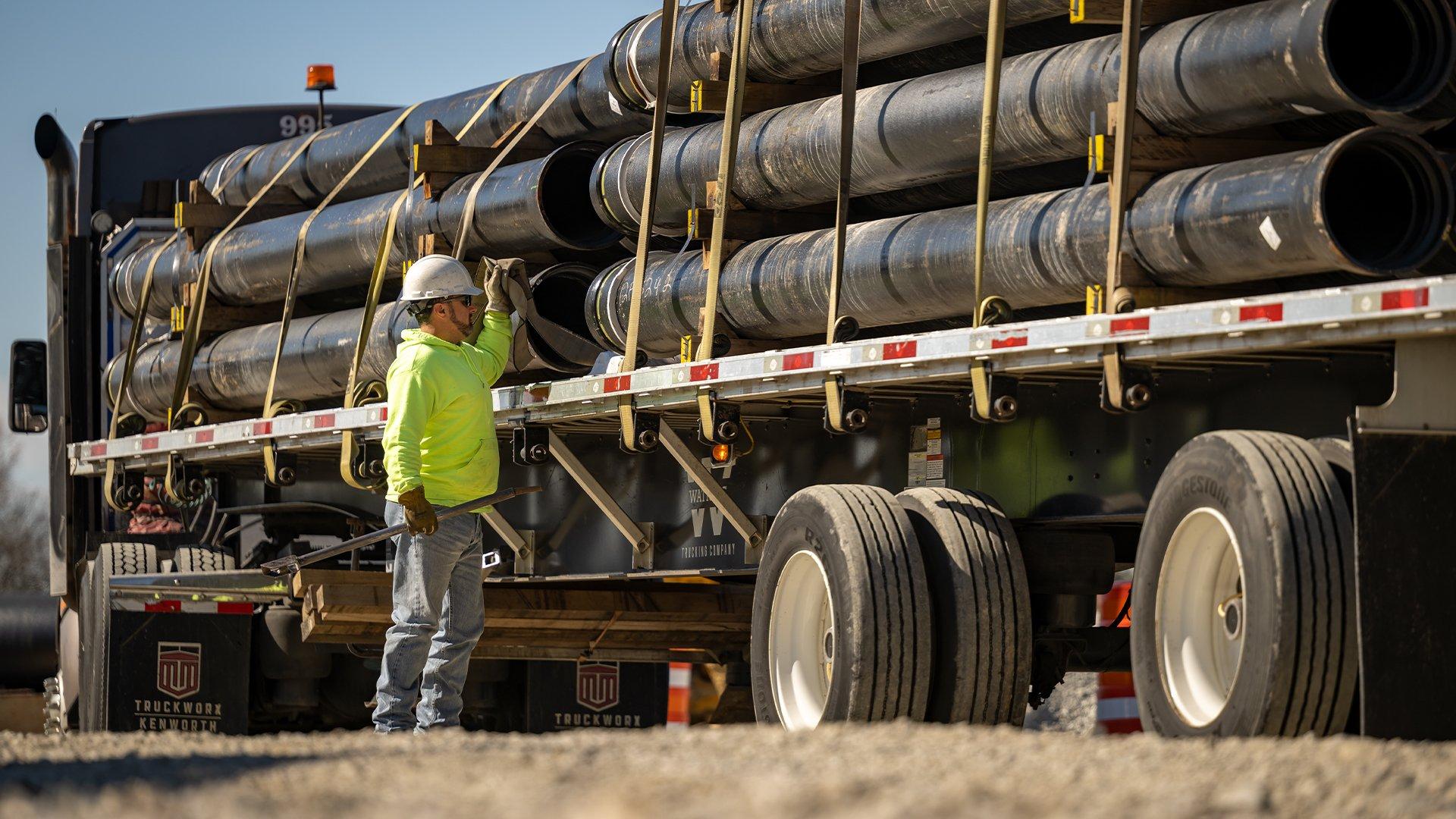 A worker wearing a hard hat checks stacks of black pipe secured on a truck flatbed.