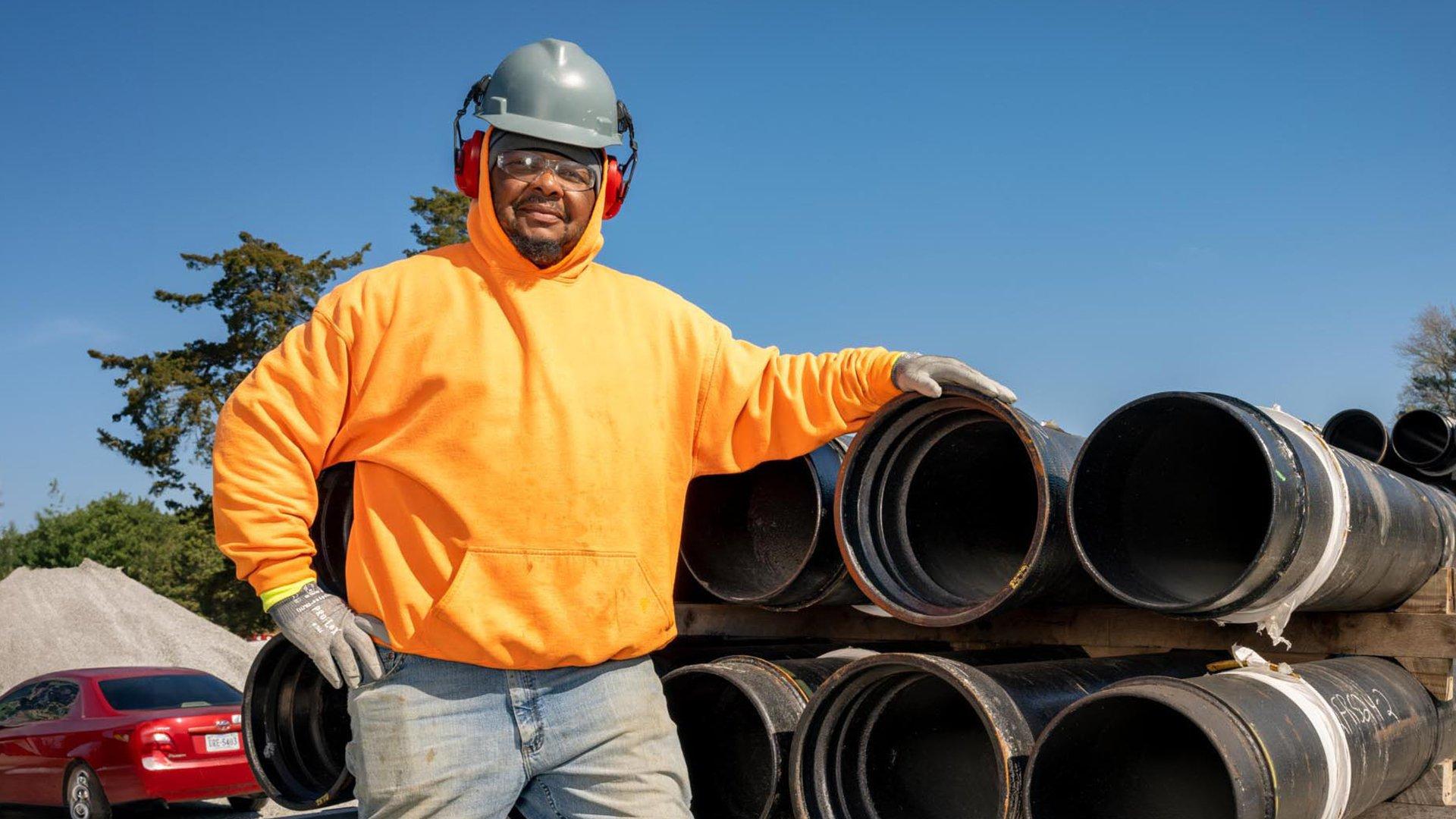 A contractor wearing a hard hat, ear protection and safety glasses stands next to a stack of pipe on a flatbed.