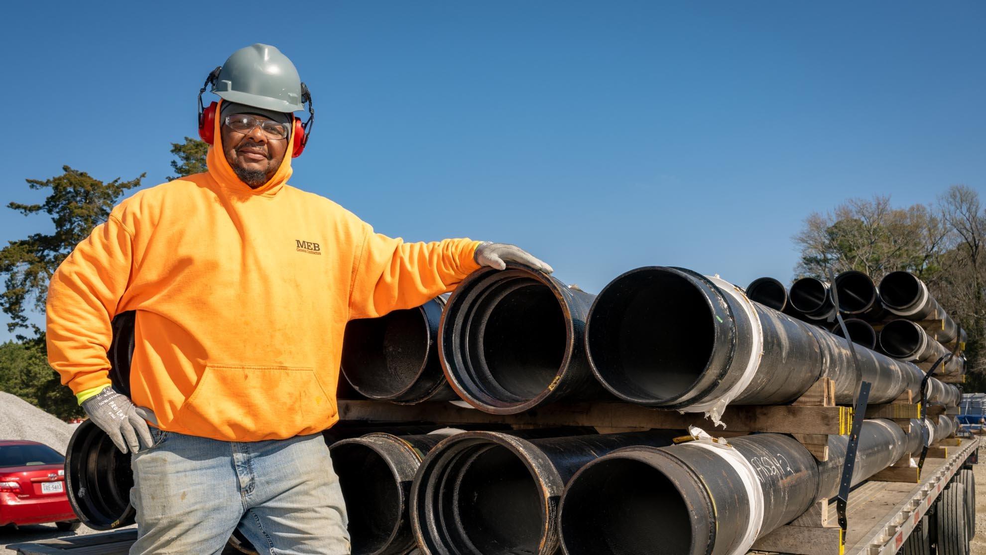 A contractor wearing a hard hat, ear protection and safety glasses stands next to a stack of pipe on a flatbed.