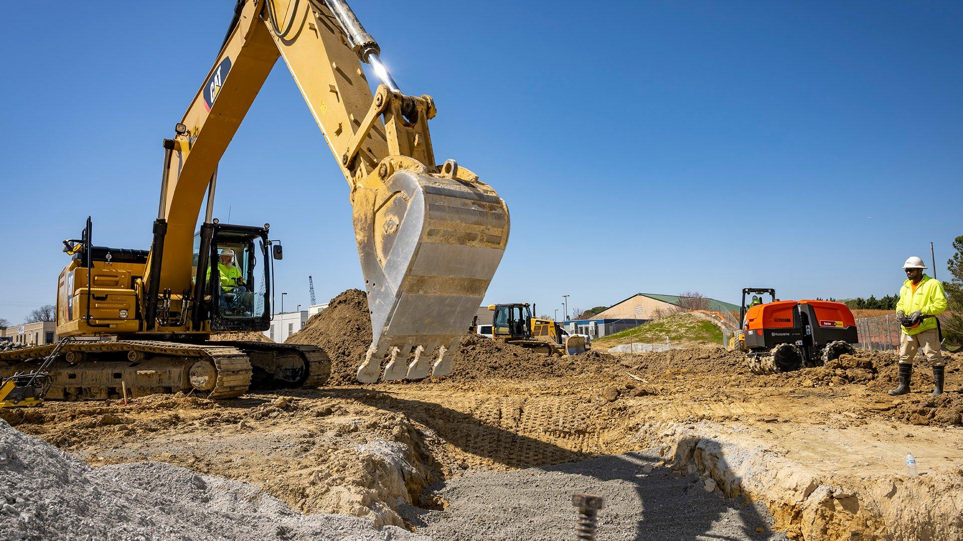 A construction worker watches while another worker operates an excavator digging a trench on a construction site.