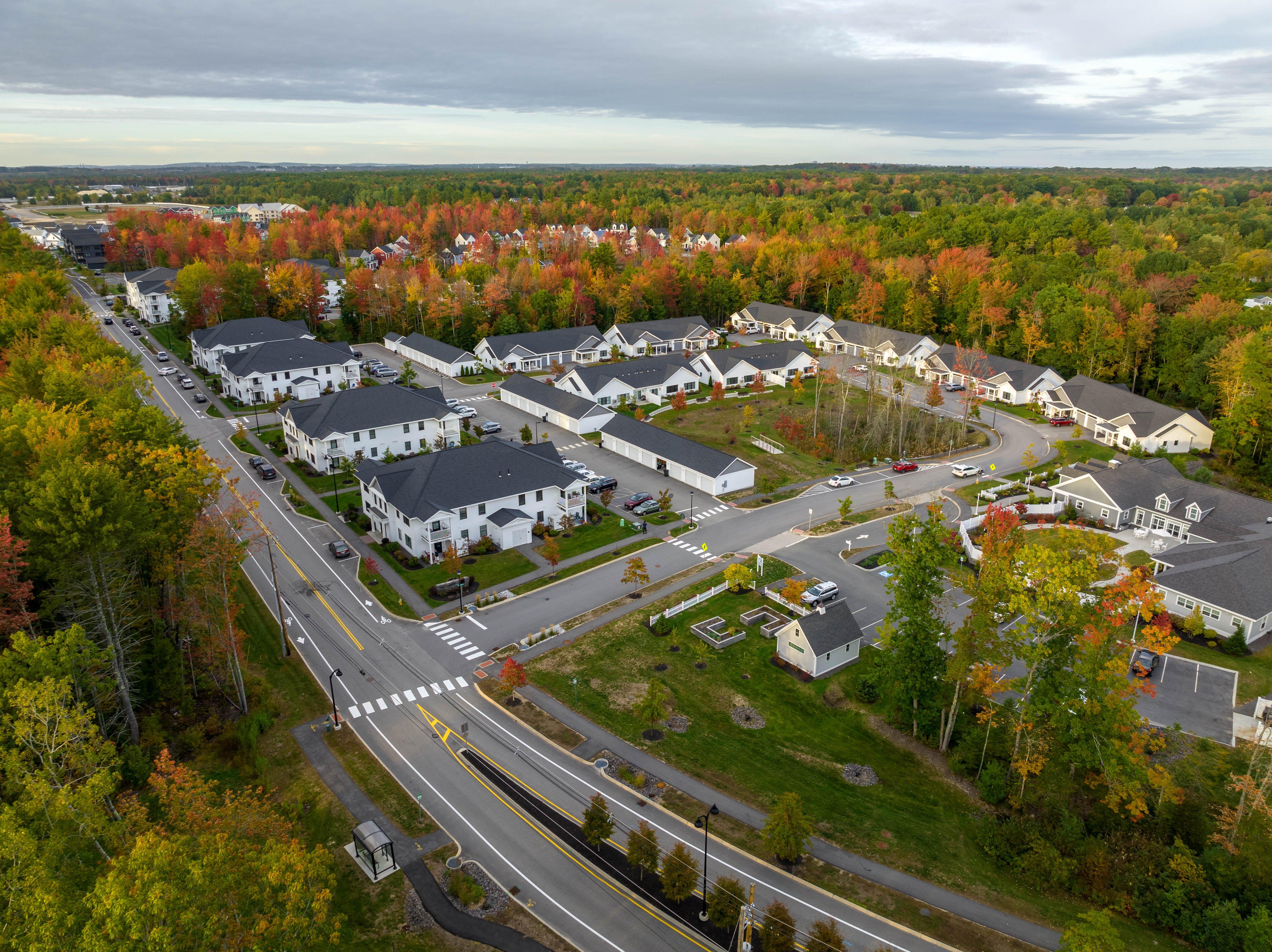 Drone view of a residential neighborhood with white homes and gray roofs, surrounded by trees.