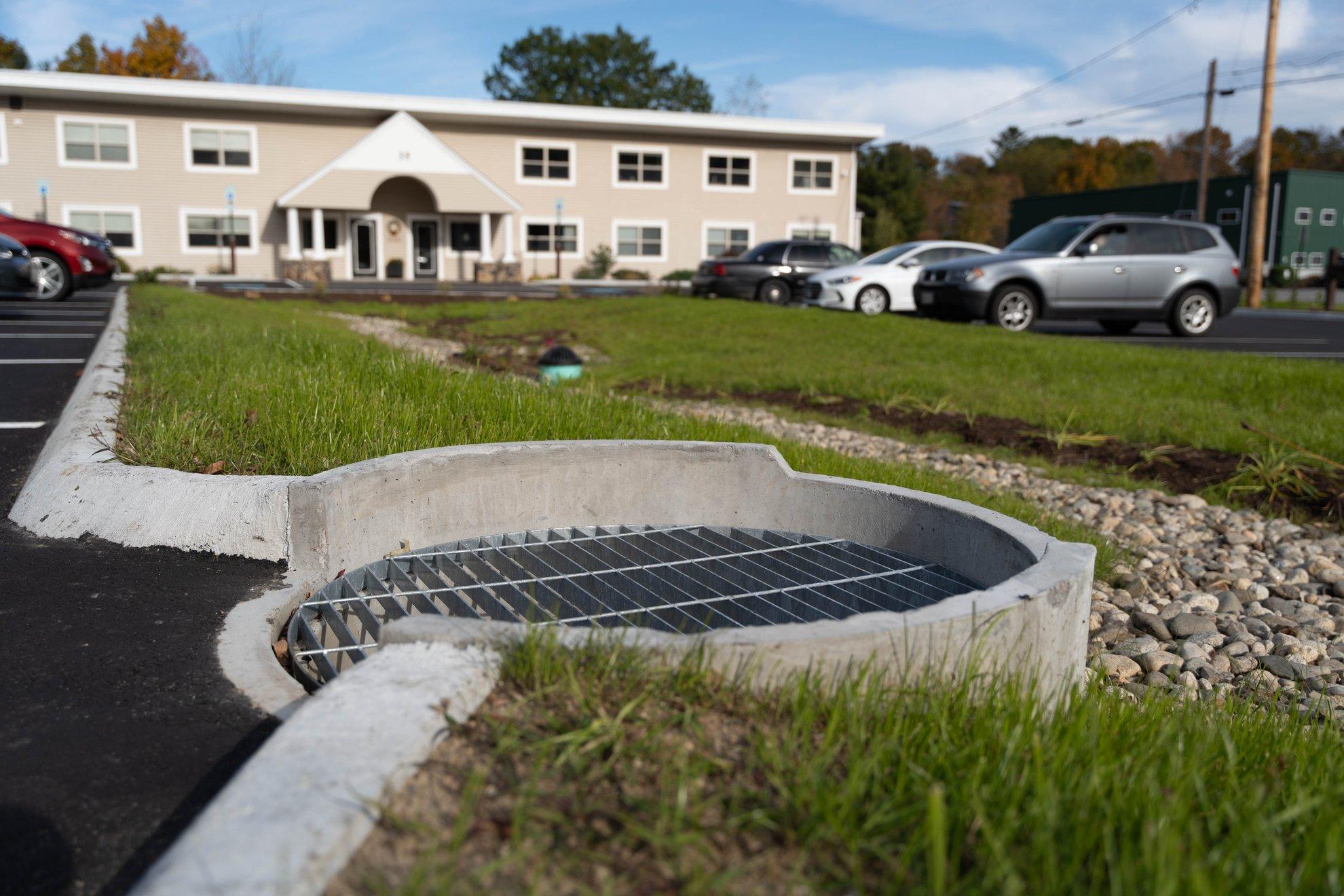 Drainage grid on a sewer tank in a grassy area, with a parking lot and office complex in the background.