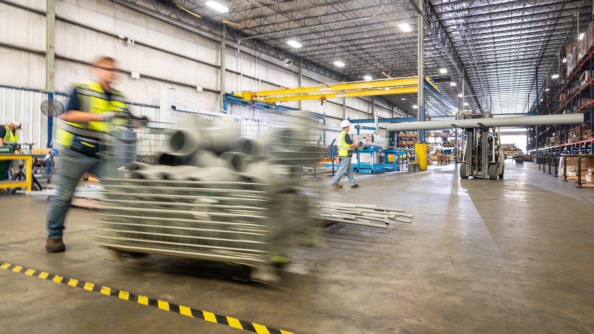 Inside of fabrication warehouse with forklift holding pipe and associate pushing pallet. 