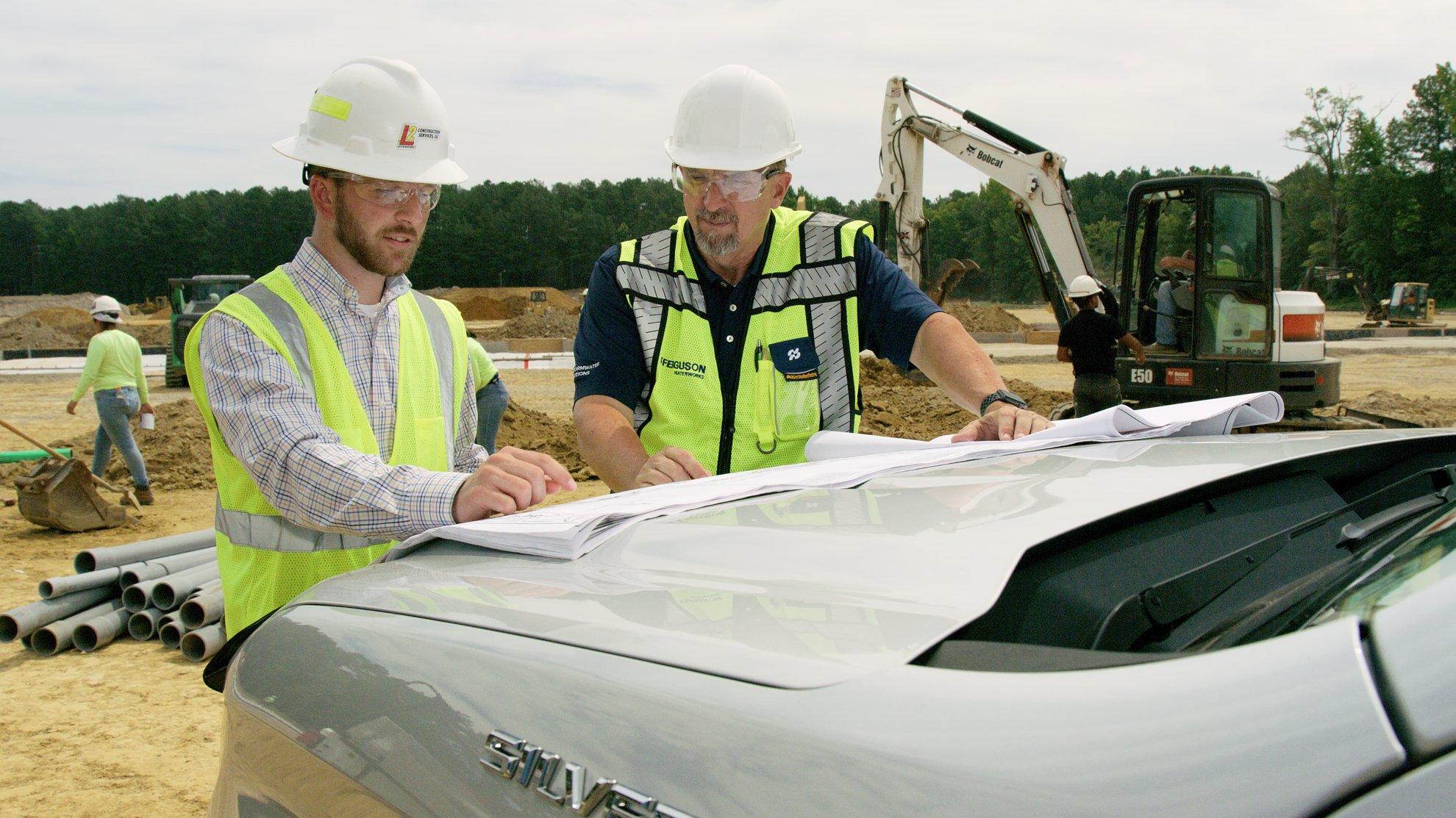 Two contractors in protective apparel review blueprints on a truck outside a commercial new build.