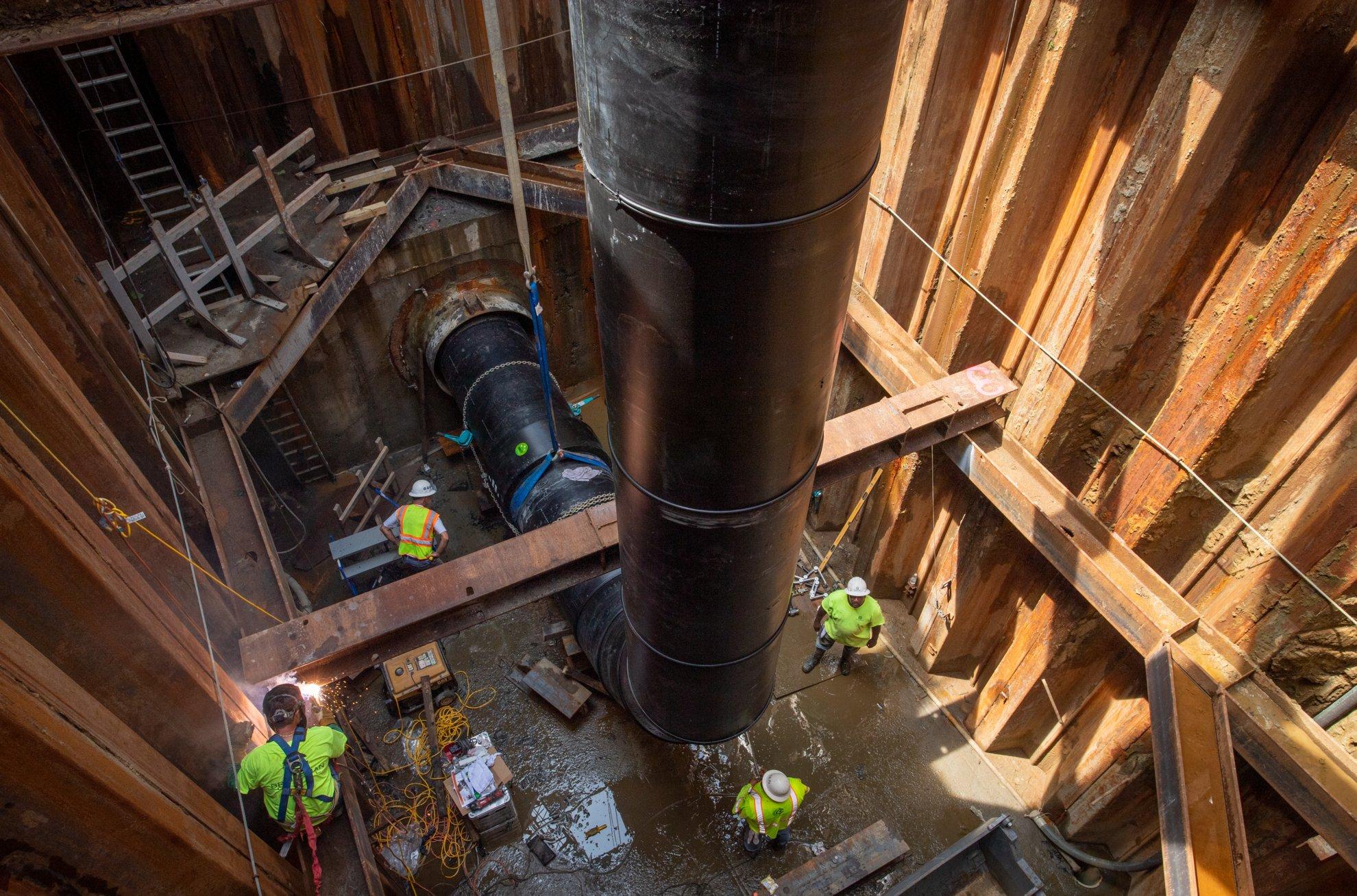 Four workers are in an underground, reinforced area around a large industrial pipe.