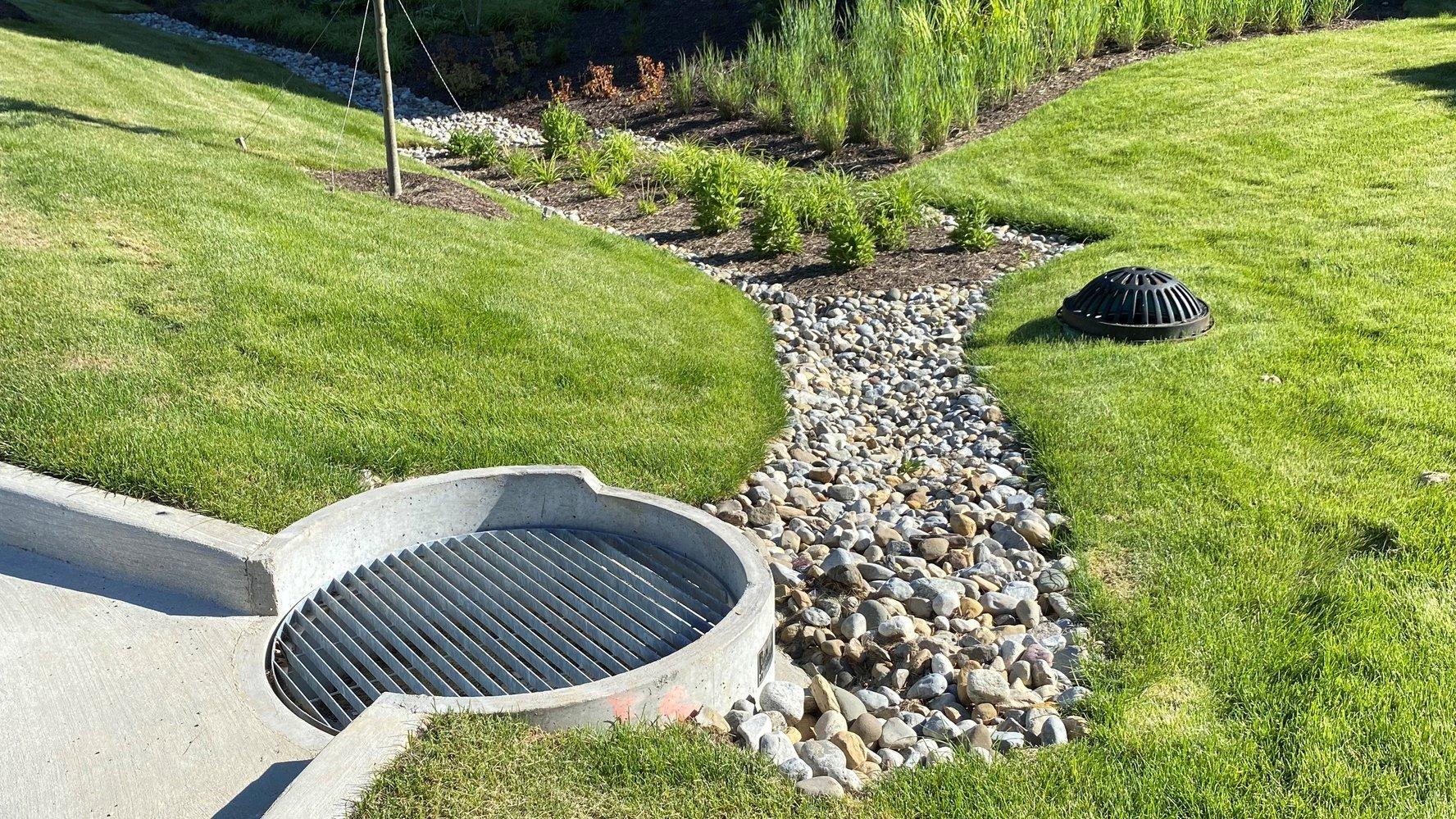 An eco-friendly drainage system, with rocks surrounded by green grass and two drainage grates.
